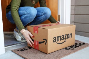 A person bends down to pick up an Amazon box delivered to a doorstep.