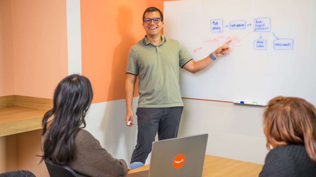 Lesson with smiling presenter pointing at a whiteboard and focused learners at table