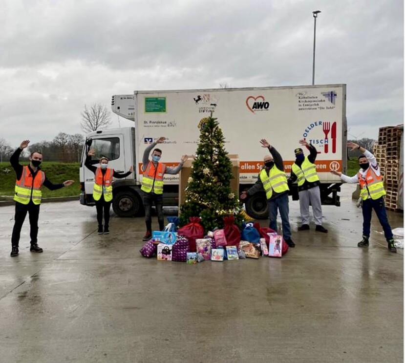Gruppenbild mit Mitarbeiter von Amazon und der Tafel vor einem Tafel-Lkw. In der Bildmitte ein Weihnachtsbaum, unter dem Geschenke liegen.