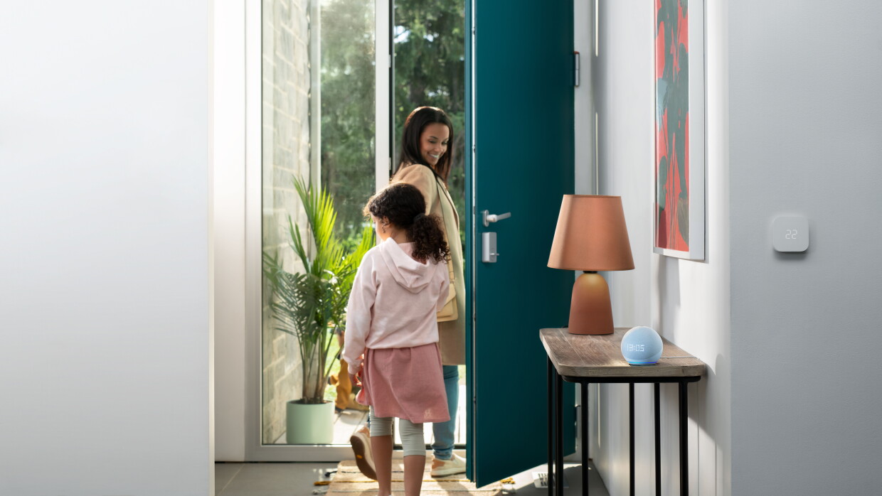 Una imagen de una mamá y su hija saliendo por la puerta de su casa. Un dispositivo Echo Dot se encuentra en la mesa al lado de la puerta.