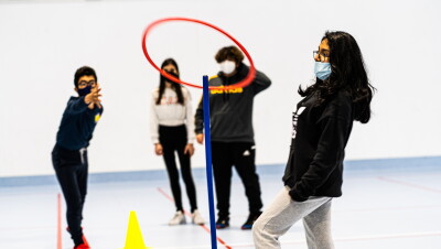 Alumnos del IESO El Melgar  (Yuncos, Toledo) en clase de educación física. Hay 4 alumnos: dos chicos y dos chicas. Una chica en primer plano con un pantalón de deporte gris y una sudadera negra Adidas- Pelo largo negro y gafas de pasta negras. En primer plano un palo vertical azul y un aro rojo a punto de entrar. En segundo plano los otros tres protagonistas. El chico vestido todo negro de deporte y con unas zapatillas rojas. Una chica con el pelo largo, unos pnatalones negros y sudadera gris y otros chico vestido todo de negro. Están en una pista de hacer deporte con el suelo azul y la pared blanca. 