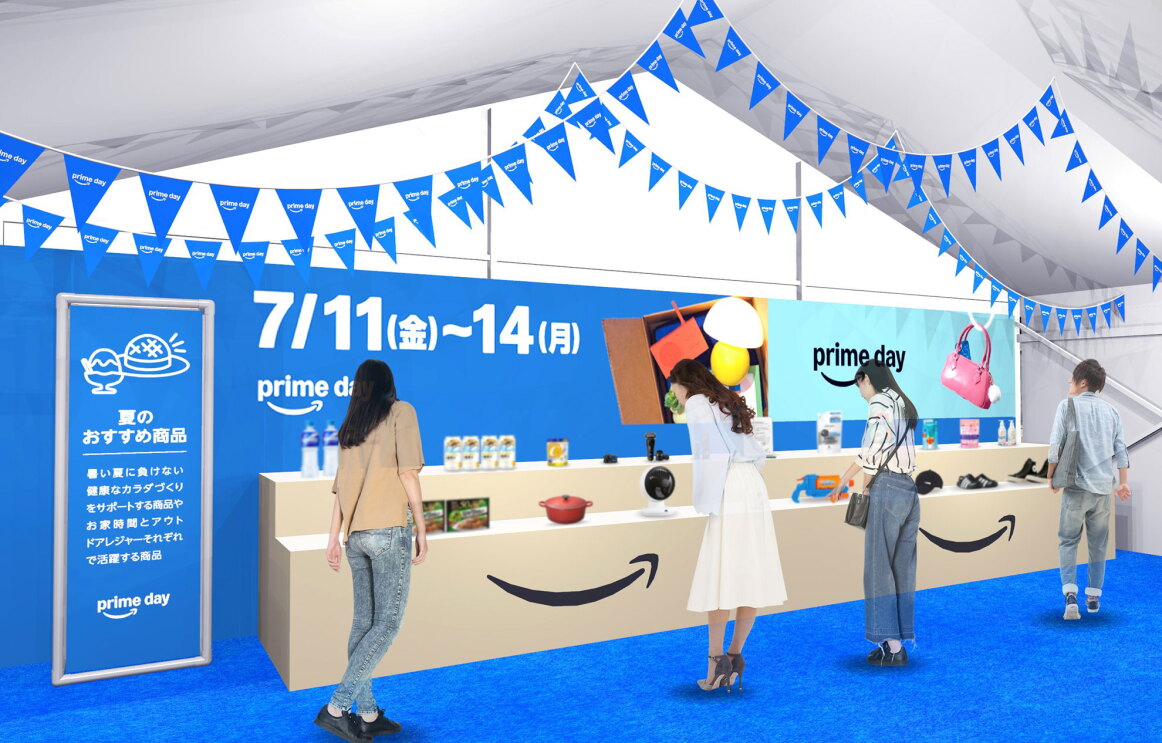 白い仮設テント内が青の床や壁、青い三画旗で装飾されている。Amazonのロゴが大きく入ったベージュの展示台にさまざまな商品が展示されている。青い壁には「7/11(金)~7/14(月)」の日程が白字で書かれている。商品展示を見て回る人々。