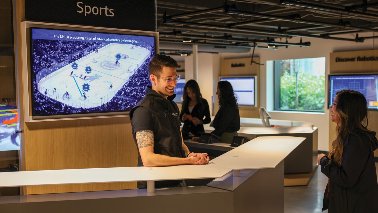 A man stands at a a counter and engages with a woman on the other side of it, inside the new AWS Skills Center in Seattle, Washington. 