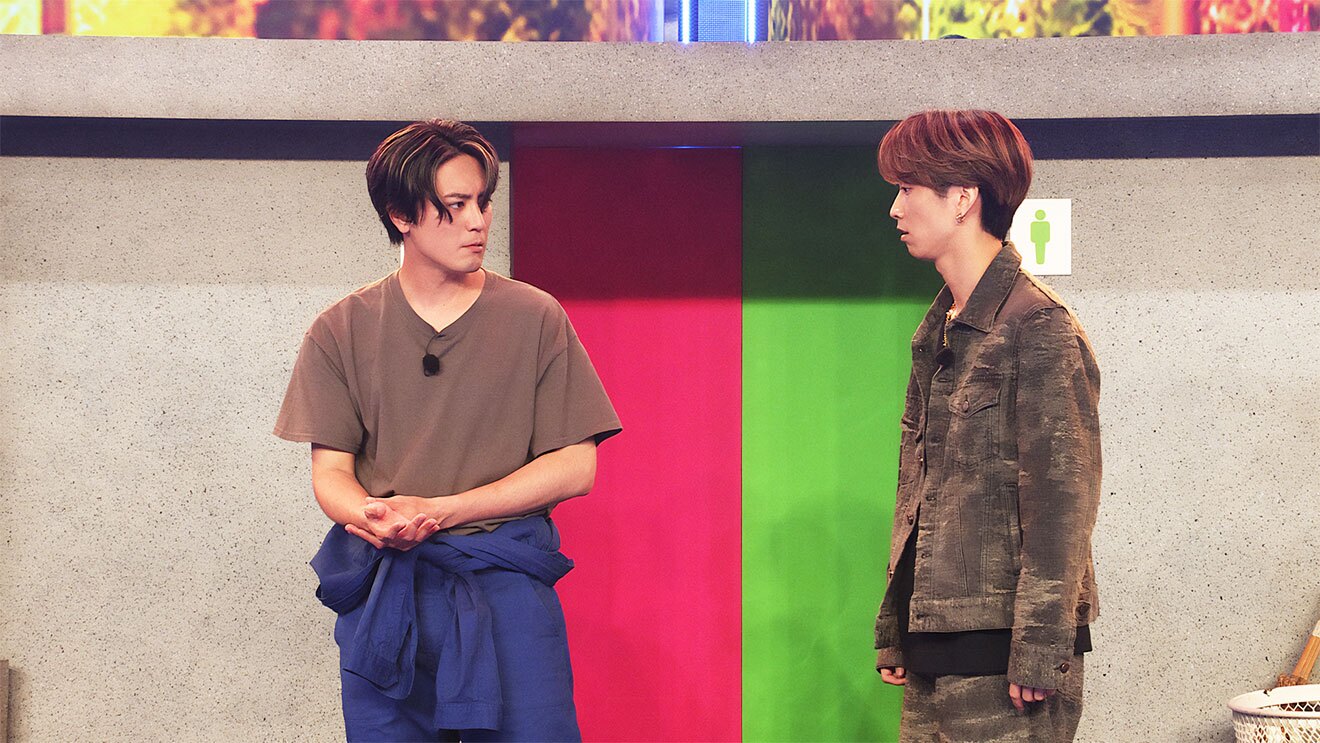 公共トイレを模したセット、ベージュグレーの壁の一部がピンクと黄緑に塗られている。その前に立つ2人の男性。左側は茶色いTシャツ、紺色の作業着を腰で巻いている。右側は茶色のセットアップ。