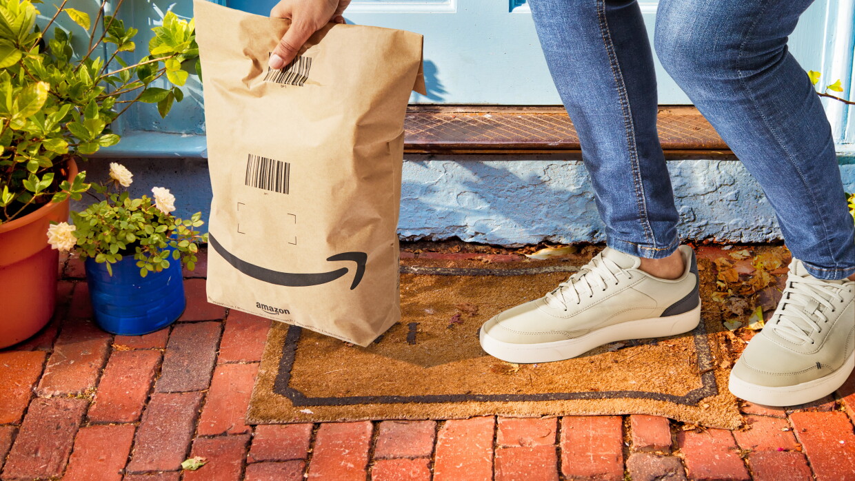 En el portal de una casa aparecen las piernas y la mano de una persona recogiendo una bolsa de papel de Amazon. 