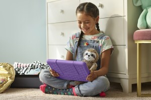 An image of a child sitting down, looking at a purple tablet and smiling. The tablet is an Amazon Fire HD 10 Kids Edition.