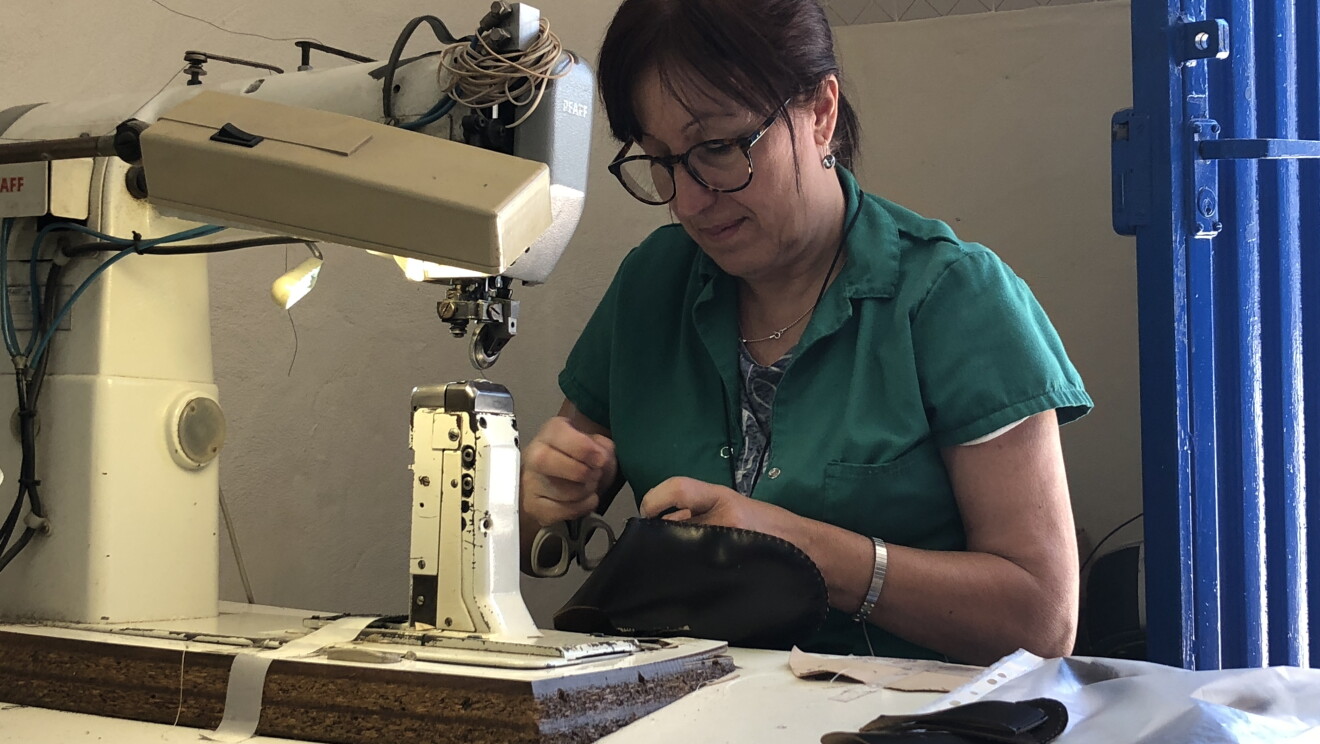 Mujer de mediana edad trabajando con una máquina de coser zaoatis, Viste una bata de color azul y en la mano tiene unas tijeras. 