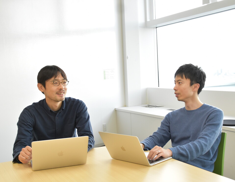 白い壁の室内で、ベージュ木目のデスクに向かう2人の男性。共に銀色のノートパソコンを開いている。
