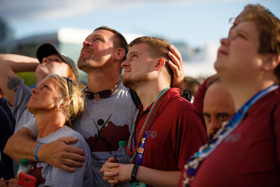 Special Olympics attendees are watching the closing ceremony festivities. In the photo, attendees are looking upward. A man embraces a woman and a younger man, as they all look skyward.
