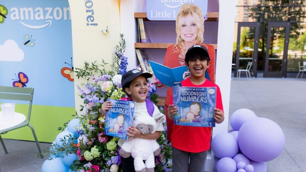 Two children holding 'Goodnight Numbers' books at an Amazon literacy event with Dolly Parton cutout