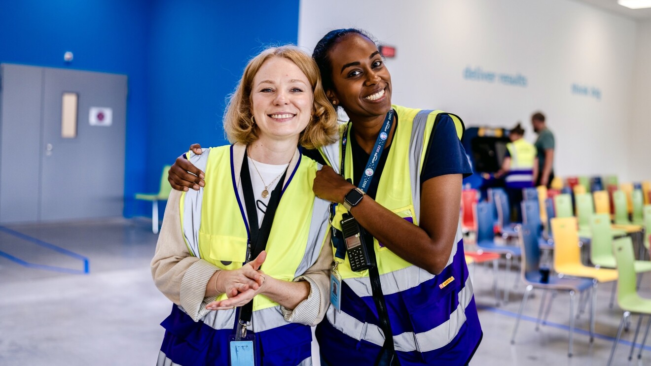 Zwei junge Amazon Kolleginnen aus Kaiserslautern mit Sicherheitswesten halten sich lächelnd im Arm: Die eine ist blond und hellhäutig, die andere ist dunkelhäutig mit dunklen Haaren.