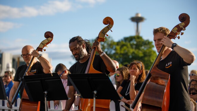 三个成员的亚马逊交响乐团演奏大提琴。每个穿着黑色t恤,站在一个乐谱架。背后的音乐家,一群与会者是可见的。