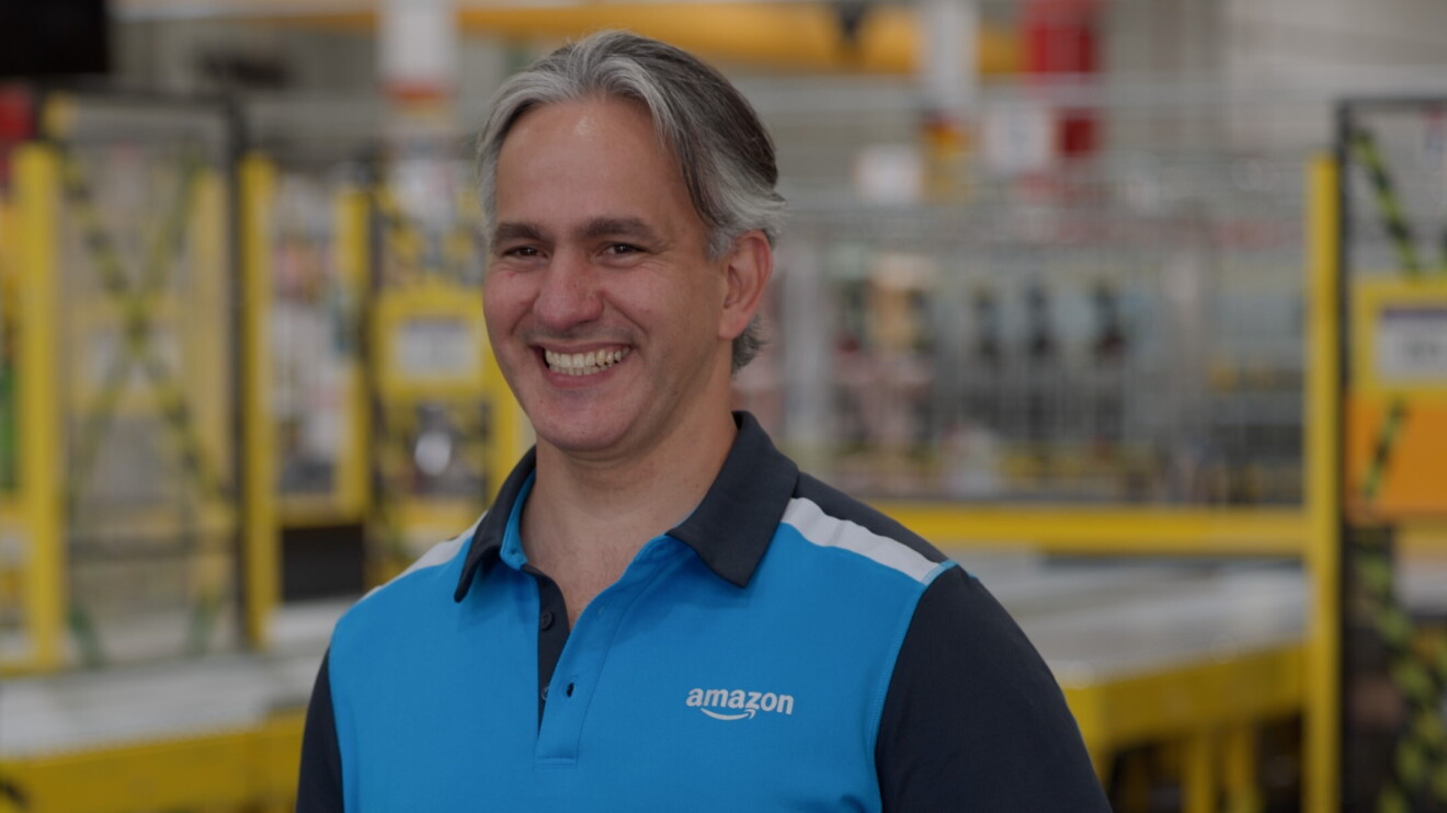 Jonathan de Dolinski - Becerra Services, S.L. mirando a cámara con una sonrisa. Lleva el pelo corto canoso y lleva un jersey de colores azules con el logo y las letras Amazon de color blanco. De fondo se ven estanterías de Amazon. 
