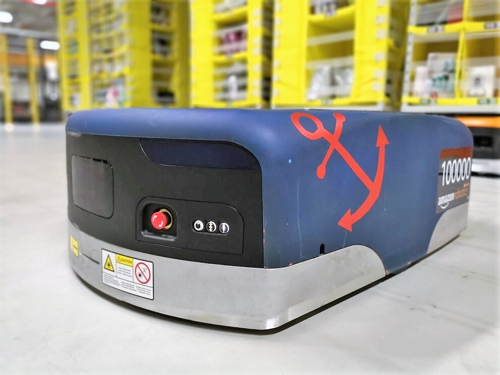 Auf dem Boden einer Lagerhalle sieht man einen blauen Amazon Transportroboter. Auf der rechten Seite wurde ein roter Anker aufgeklebt und ebenfalls rechts steht die kennzeichnung "100000 amazon robotics".