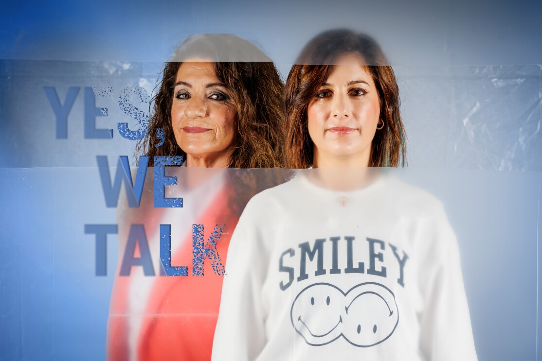 Dolores y Pilar en un plano hasta la cintura. Dolores va con una americana rosa y debajo una camisa blanca. Pilar lleva una sudadera blanca con dos emoticonos con cara sonriente y el texto Smiley. En el lado izquierdo está el lema de la campaña Yes, we talk, debajo rompiendo el silencio. El texto es de color azul y el plástico también tiene el mismo color que se va degradando hasta convertir en blanco en el centro de la imagen. Las protagonistas miran a cámara. Hay un plástico que está cortado para que se pueda ver su cara nítida.