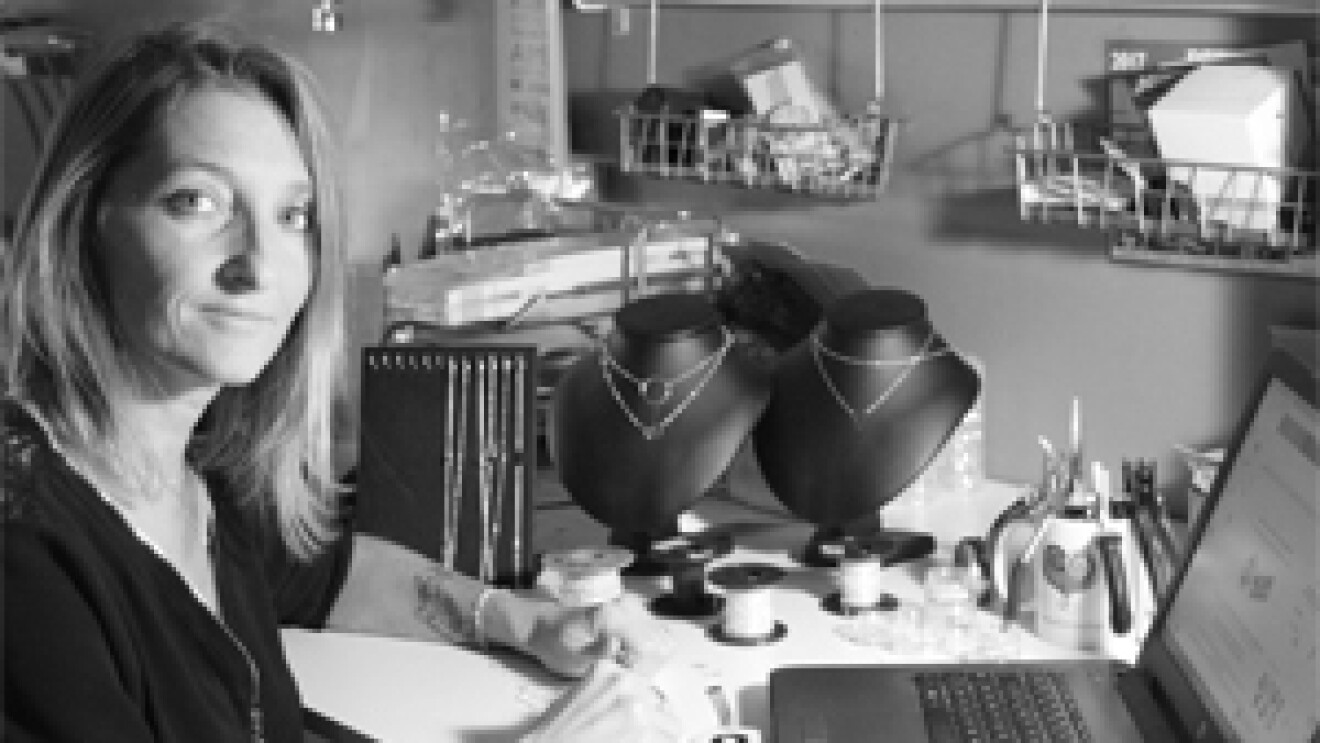 La fondatrice d'EmmaFashionNow se tient devant une table où se trouvent des bijoux finis ou en cours de création ainsi que son ordinateur. C'est une photo noir et blanc.