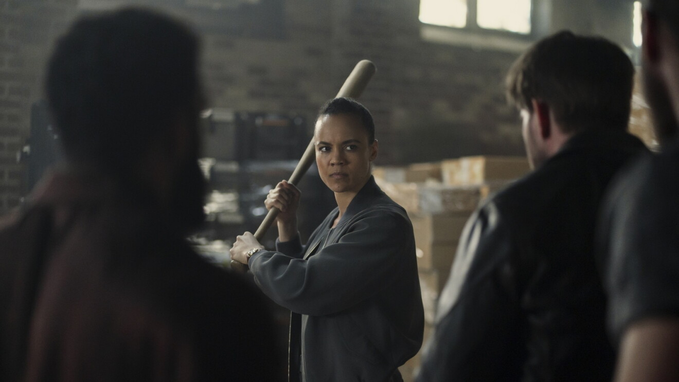 Still from 'Neagley' of woman in dark jacket holding baseball bat in defensive stance while confronting two people in warehouse