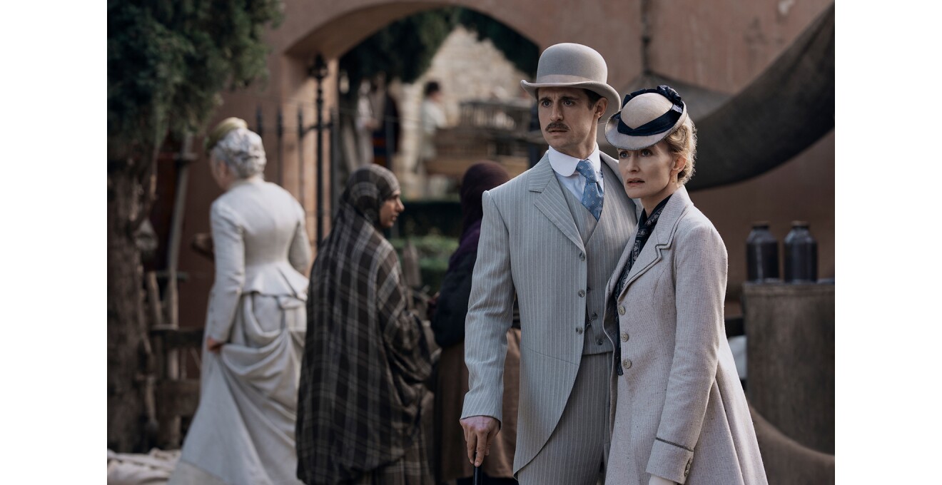 日中の街中に並んで立つ男女。共に西洋の正装で帽子をかぶり、男性はステッキを使用。背後には、女性と同じような洋風ドレスの女性が1人、異なる文化圏の民族衣装を着た2人の人物。