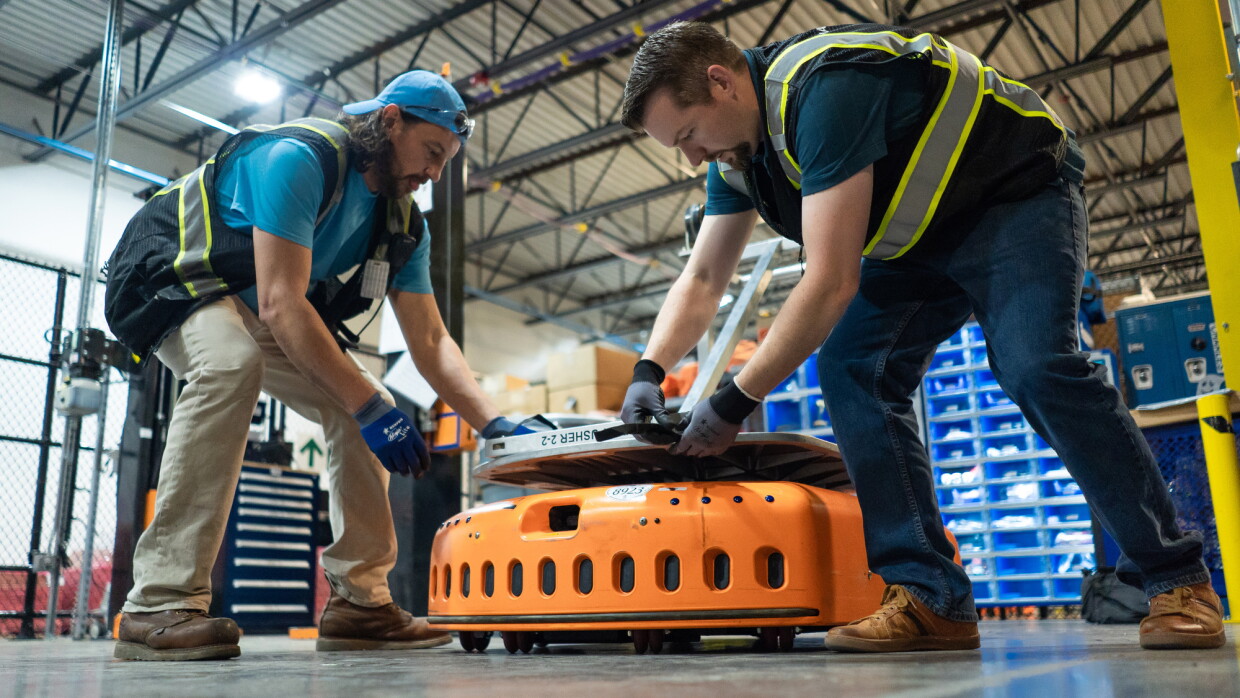 Two men in a warehouse space reach down to secure a silver-colored object to an orange, wheeled piece of machinery.