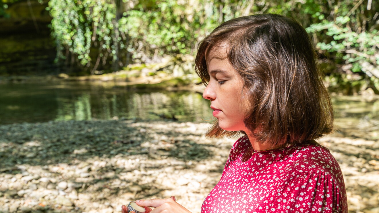 María Gómez Iribarnegaray con el  bálsamo labial de La cosmética de María.  María esñta embarazada y lleva un vestido rosa y  blanco de manga larga y cuello redondo. De fondo un río con piedras y verde.  En la mano tiene el recipiente del bálsamos y un anillo. 