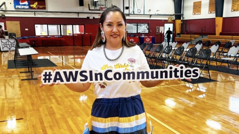 Indigenous woman holds up a sign that reads 'AWS In Communities' in a high school gym.