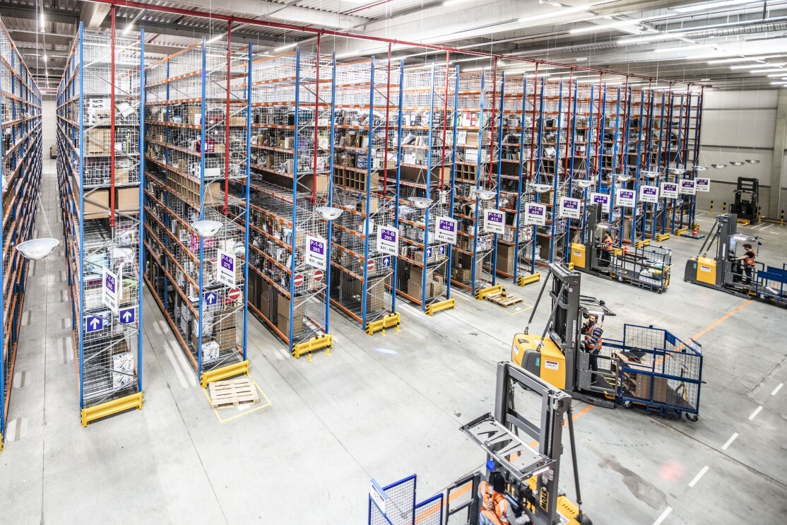 Rund teilweise gefüllte Hochregallager sind zu sehen, vor ihnen fahren vier Kommissionierfahrzeuge.