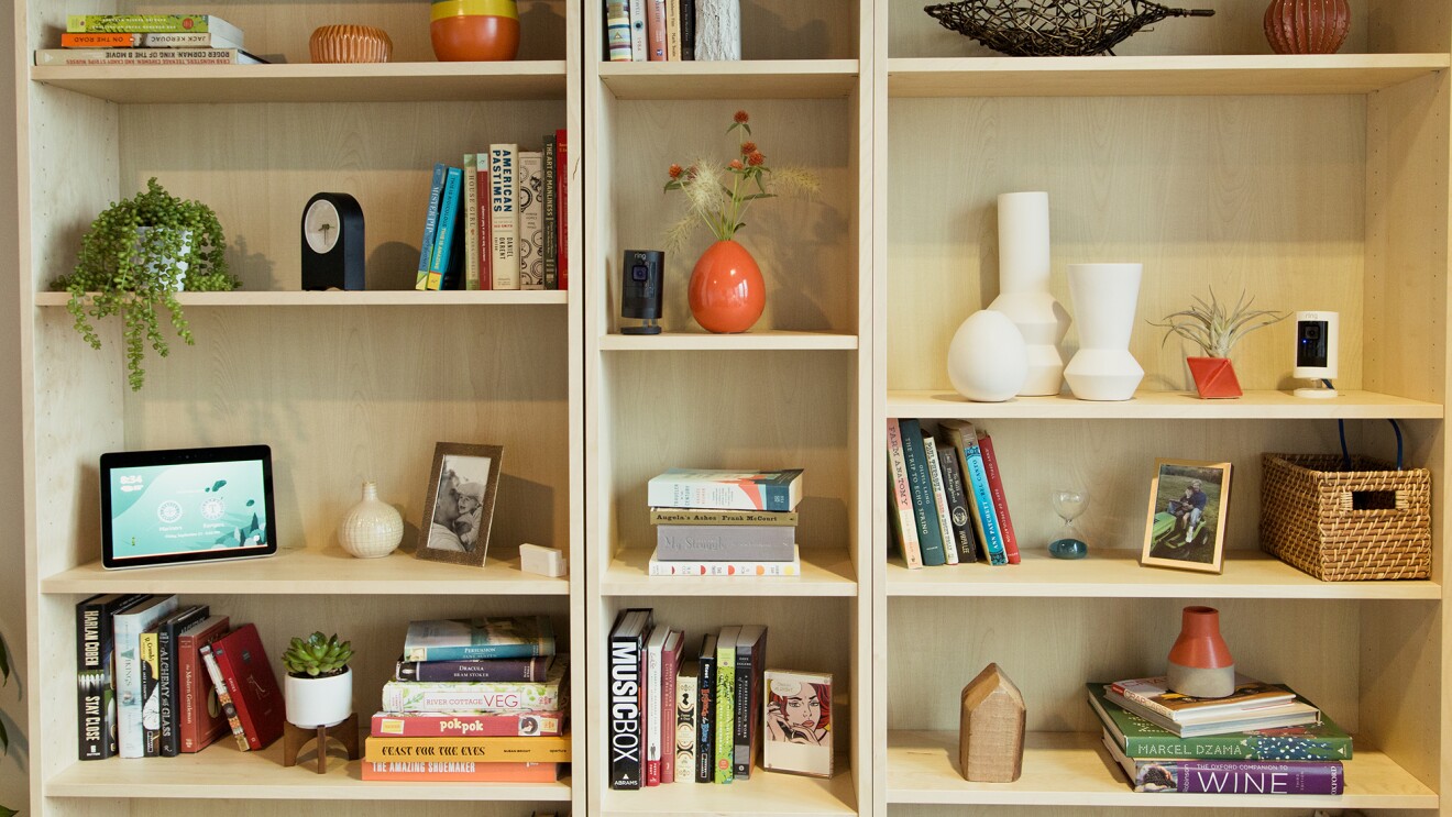 A bookshelf with books, decor, vases and baskets, clock, Echo Show, and Ring camera, illustrating a Smart Home setting.