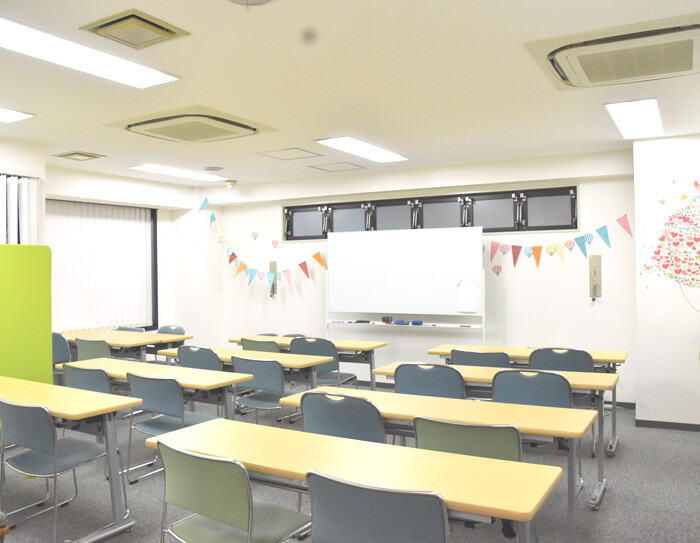白い壁、白い天井の教室。前方の壁にホワイトボード。複数の長机に椅子が2脚ずつ並んでいる。