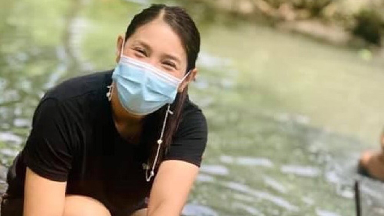 An image of a woman working near a river with a coconut in front of her. She has a surgical mask on.