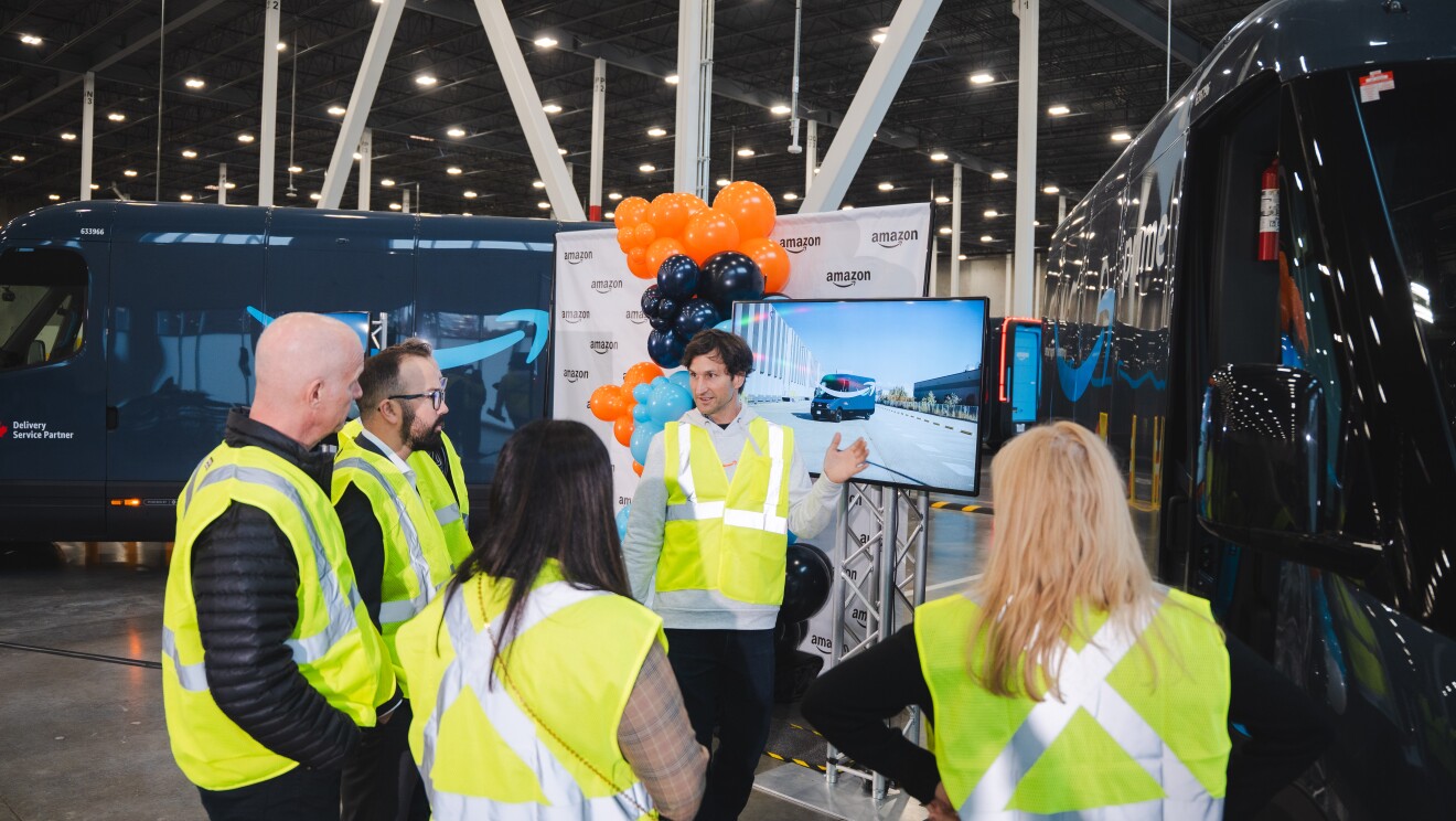 Image of a man speaking in front of a screen displaying an Amazon x Rivian delivery van to people in yellow vests