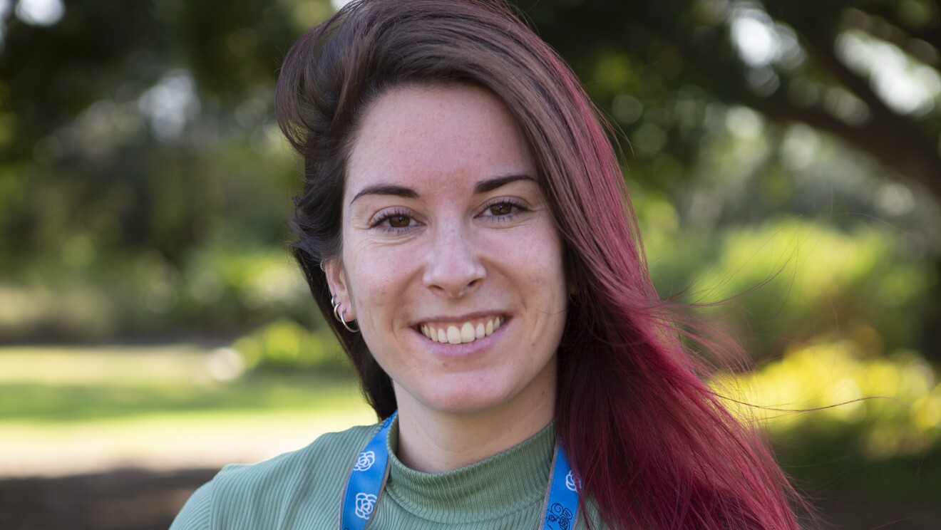 Lucía Galve es la protagonista de la foto. Mira a cámara y sonríe con los brazos detrás, en la espalda. Lleva el pelo largo, de color castaño pero con las puntas rojas y suelto. Va con unos pantalones de color negro, un jersey de punto de color verde. La foto es exterior y de fondo, difuminado unos árboles y una campo con césped. 