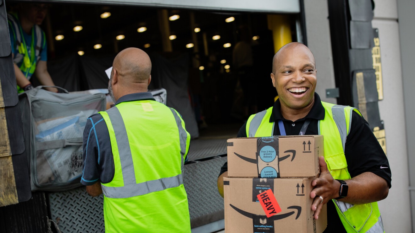 A smiling man holds boxes while people in the background load a truck. 