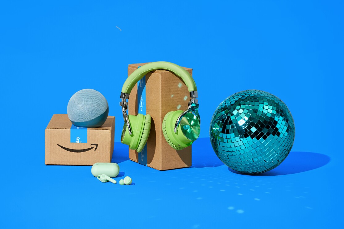 Fotomontaje, sobre un fondo azul, dos cajas de Aamzon, una con dos dispositivos Amazon apoyados, la caja grande con unos auriculares verdes y al lado derecho una bola de fiestas de discoteca.