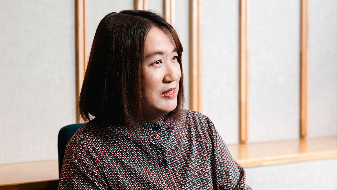 ベージュの壁に、木製の仕切りが複数配置された明るい室内で、赤と紺の細かい幾何学模様のワンピースを着た女性が椅子に座って話している