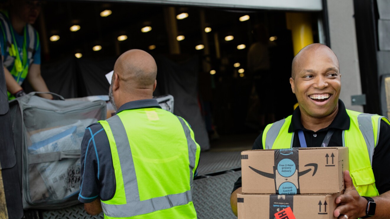 A smiling man holds boxes while people in the background load a truck.