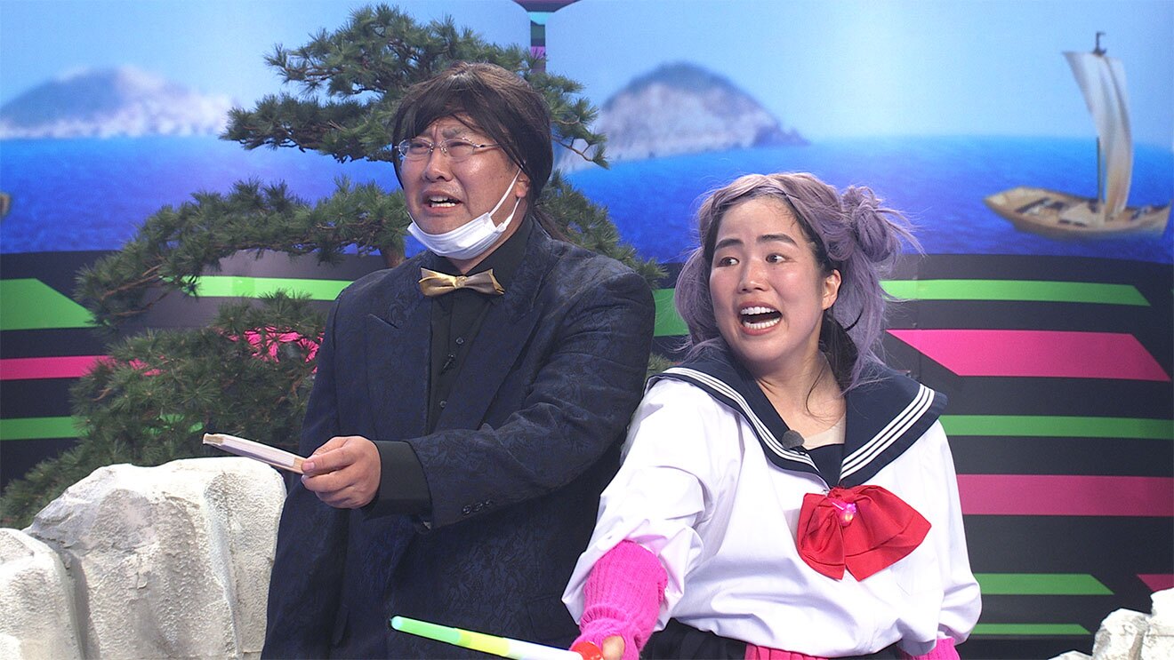 海の風景画、岩と松の木が置かれたセットに立つ男女。左の男性は紺のスーツであごにマスクをかけ、右はセーラー服で紫のかつらをかぶった女性。2人とも手に持ったものを前に突き出す。