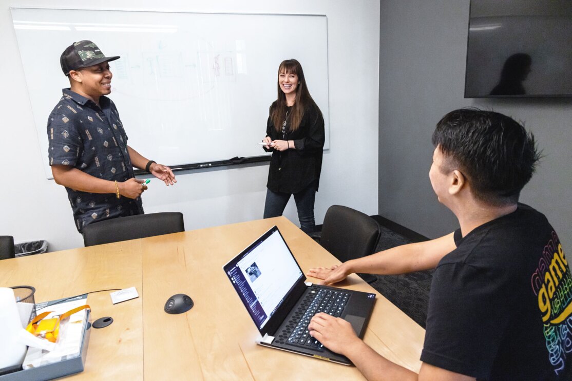 An image of three people collaborating in a conference room with a laptop open and a whiteboard behind them. 