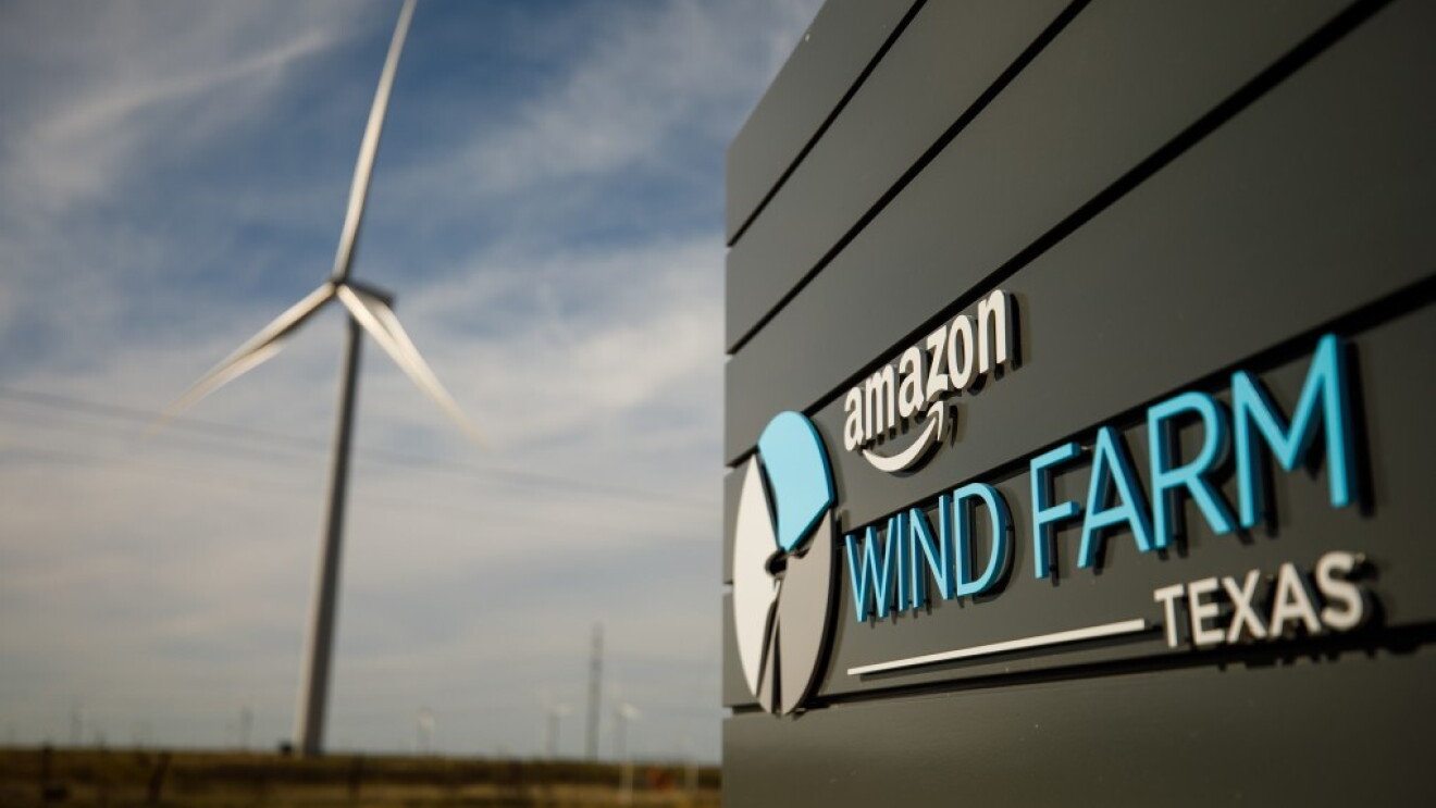 Parco eolico di Amazon in Texas. Pala eolica sullo sfondo con cielo azzurro e nuvole. Logo Amazon Windfarm Texas in primo piano, scritto in bianco e celeste. 