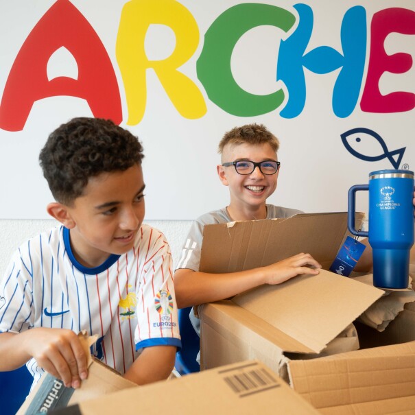 Zwei Kinder packen Pakete aus und stehen vor der Aufschrift "Arche". 