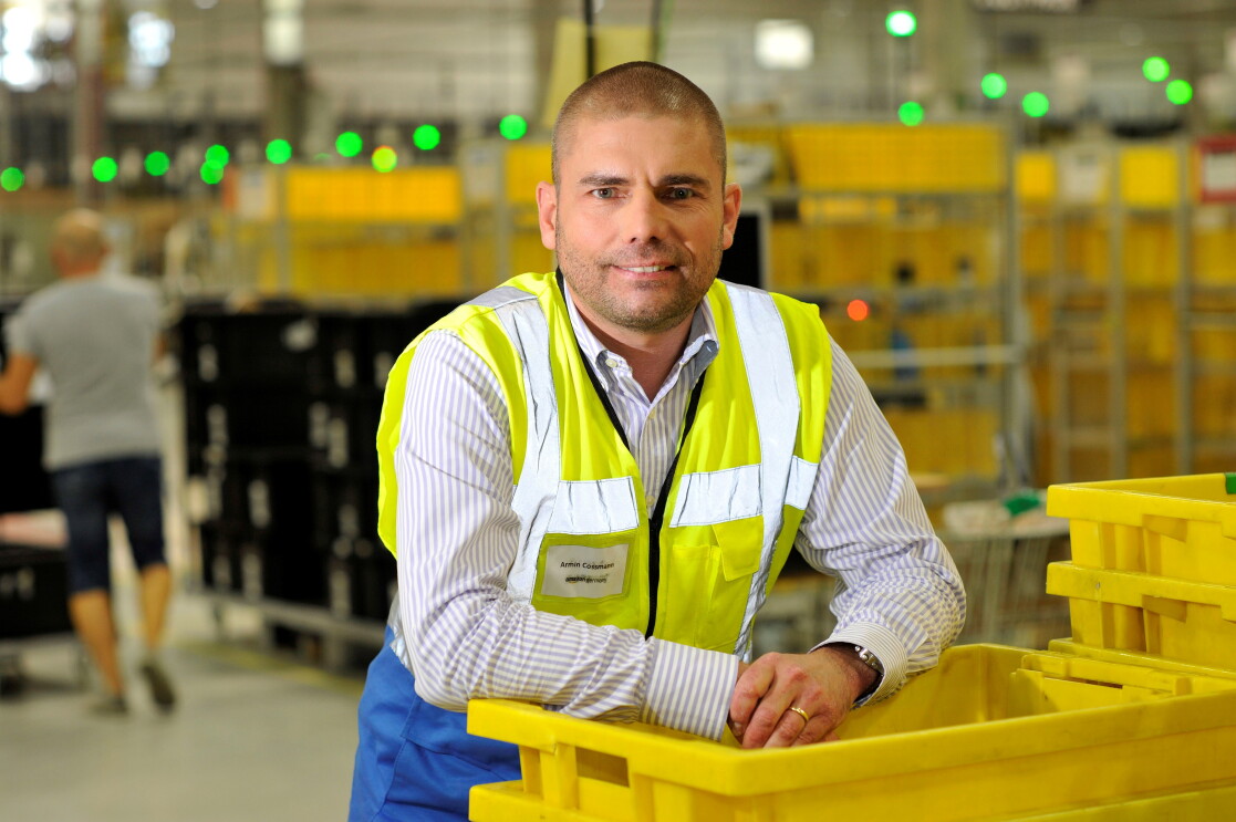 Ein Mann in gelber Sicherheitsweste lehnt sich lässig auf einem Stapler gelber Transportbehälter im Logistikzentrum