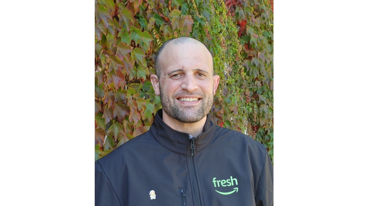 A headshot image of a man smiling for a photo in front of a wall with green and read vine leaves on it. 