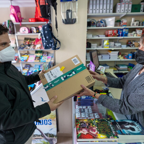 Ana Gallardo regente El Detallito, una papelería y agencia de viajes en Los Corrales, Sevilla. En la papelería Ana entrega un paquete a un cliente. Los dos llevan mascarilla. Ana está detrás del mostrados lleno de libros. En el fondo estanterías llenas de productos de papelería. Ana va con una chaqueta gris, media melena y una mascarilla negra. El cliente va con un anorak negro y una mascarilla blanca y tiene el pelo corto negro. 