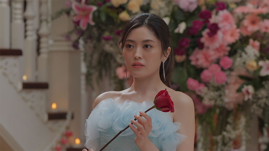 たくさんの生花が飾られた屋内に立つ女性。薄い水色のドレスを着用、体の前で一輪の赤いバラを持っている。
