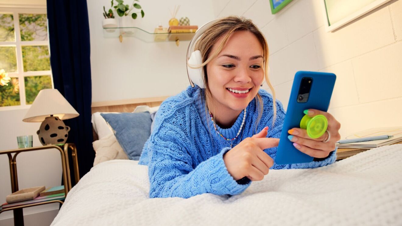 An image of a woman wearing a blue sweater and headphones reading from her phone while on her bed.