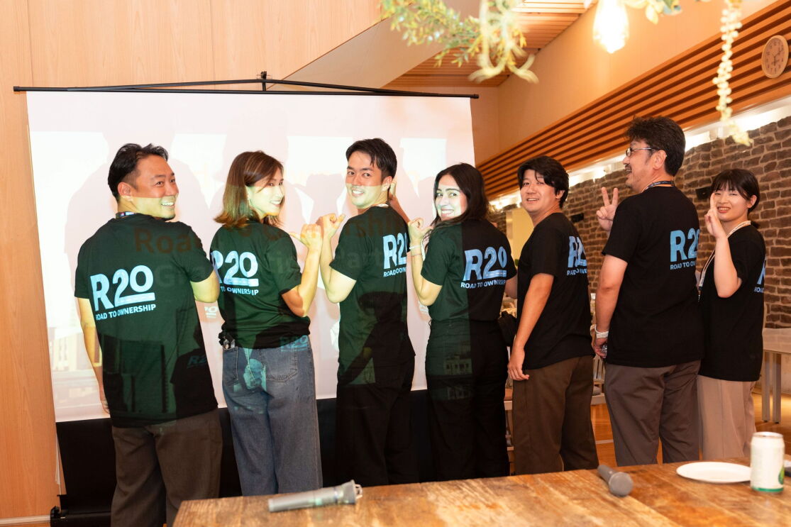 R2Oという青文字で印刷されているTシャツを着ている男性4人と女性3人が後ろに向かって並んでいる。Tシャツの後ろを見せさせている。