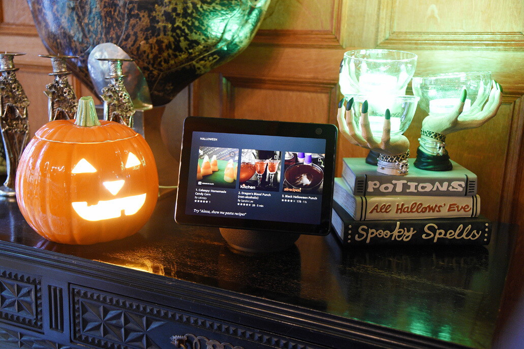 An image of an Echo Show device sitting atop a hallway table next to Halloween decorations. Halloween recipes are showed on the Echo Show screen. 