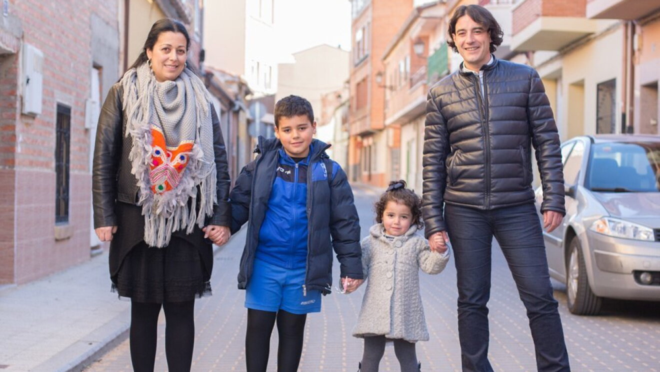 Carmelo Castro en una calle de Pozoantiguo, Zamora, acompañado de su esposa y sus dos hijos. Él viste casual yella con un vestido negro y un pañuelo gris y naranja. Él hijo va vestido para ir a entrenar a fútbol y la hija con unos leotardos y una chaqueta gris. 
