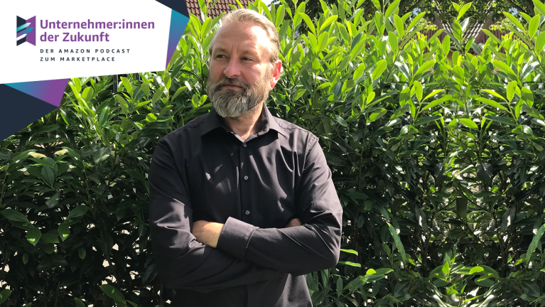 Der Gast des Unternehmer:innen der Zukunft Podcasts,  Christian Lier von Hana Lima, steht mit dunklem Hemd und verschränkten Armen im Garten vor einer grünen Hecke.