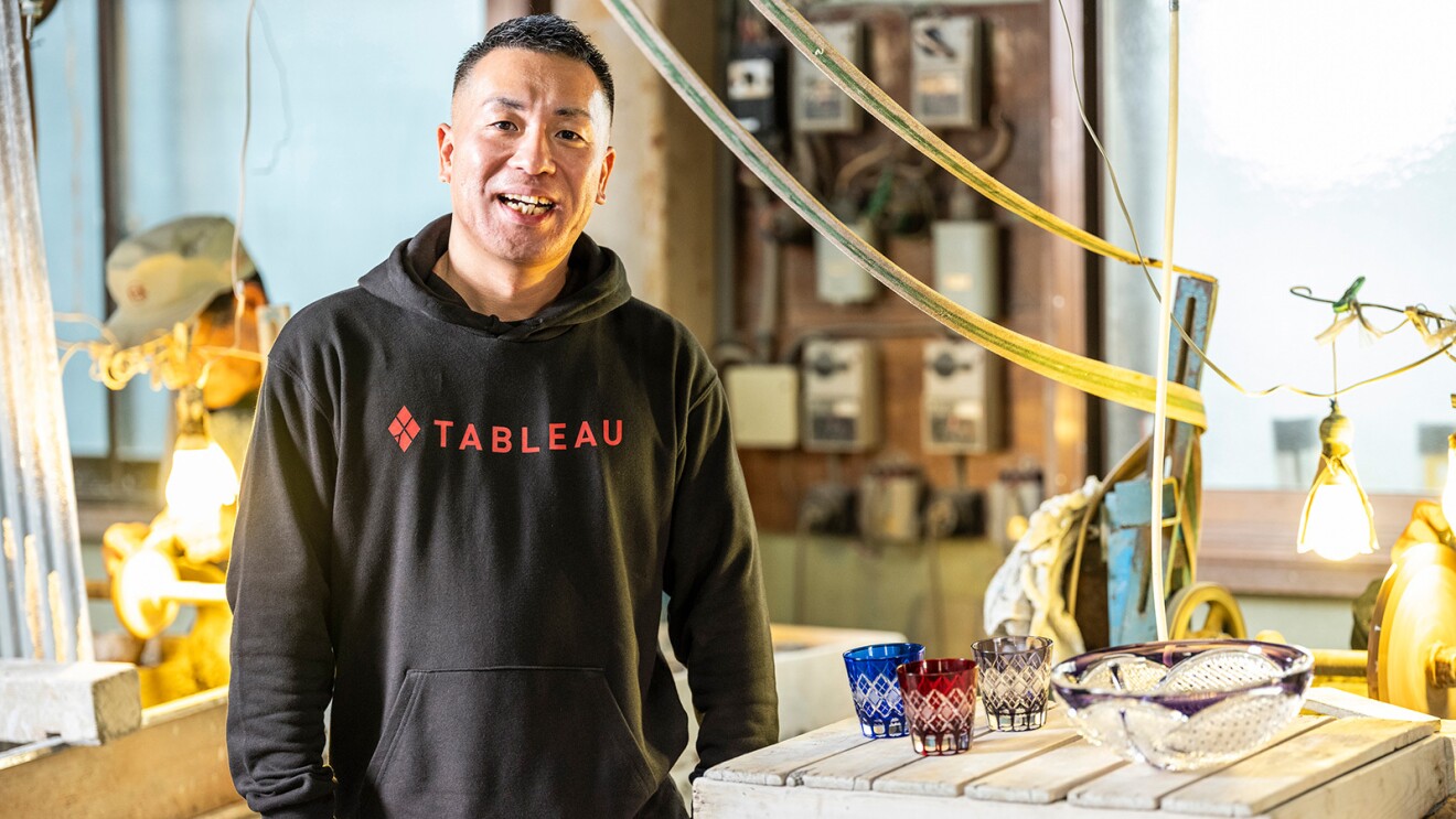 工場の中に立つ黒いパーカーを着た男性。背後で職人が作業をしている