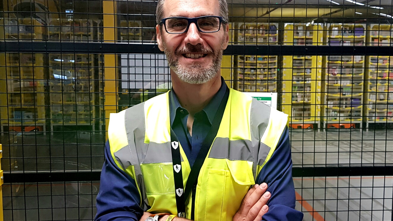 Salvatore Iorio, Country HR Manager di Amazon Italia Logistica, sorride a mezzo busto con le braccia conserte all'interno di un'area di un magazzino Amazon. Indossa degli occhiali da vista, un giubbino catarinfrangente giallo e una camicia blu.